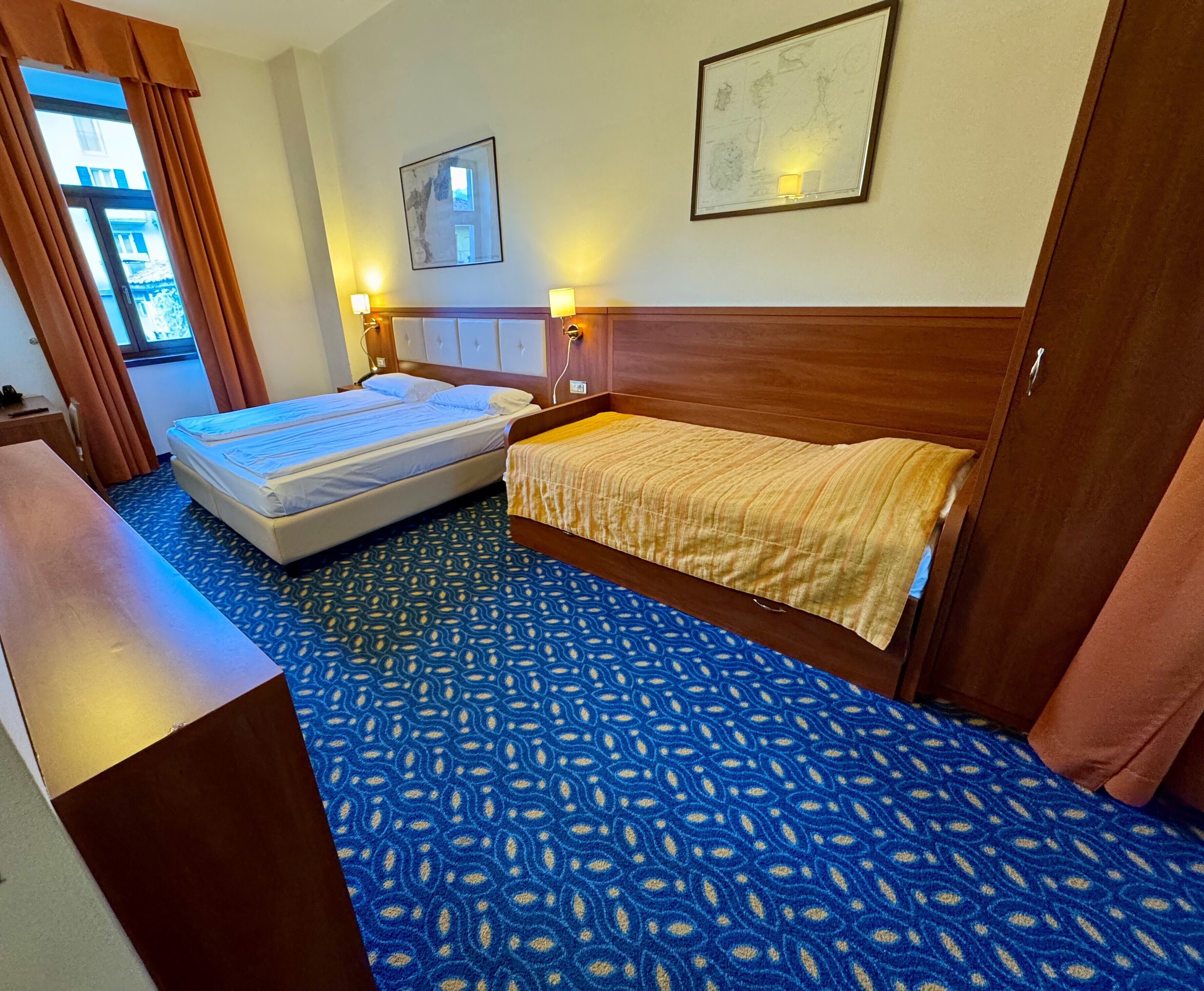 A hotel room with two beds, a wooden headboard, patterned carpet, a desk, and two wall-mounted lamps.