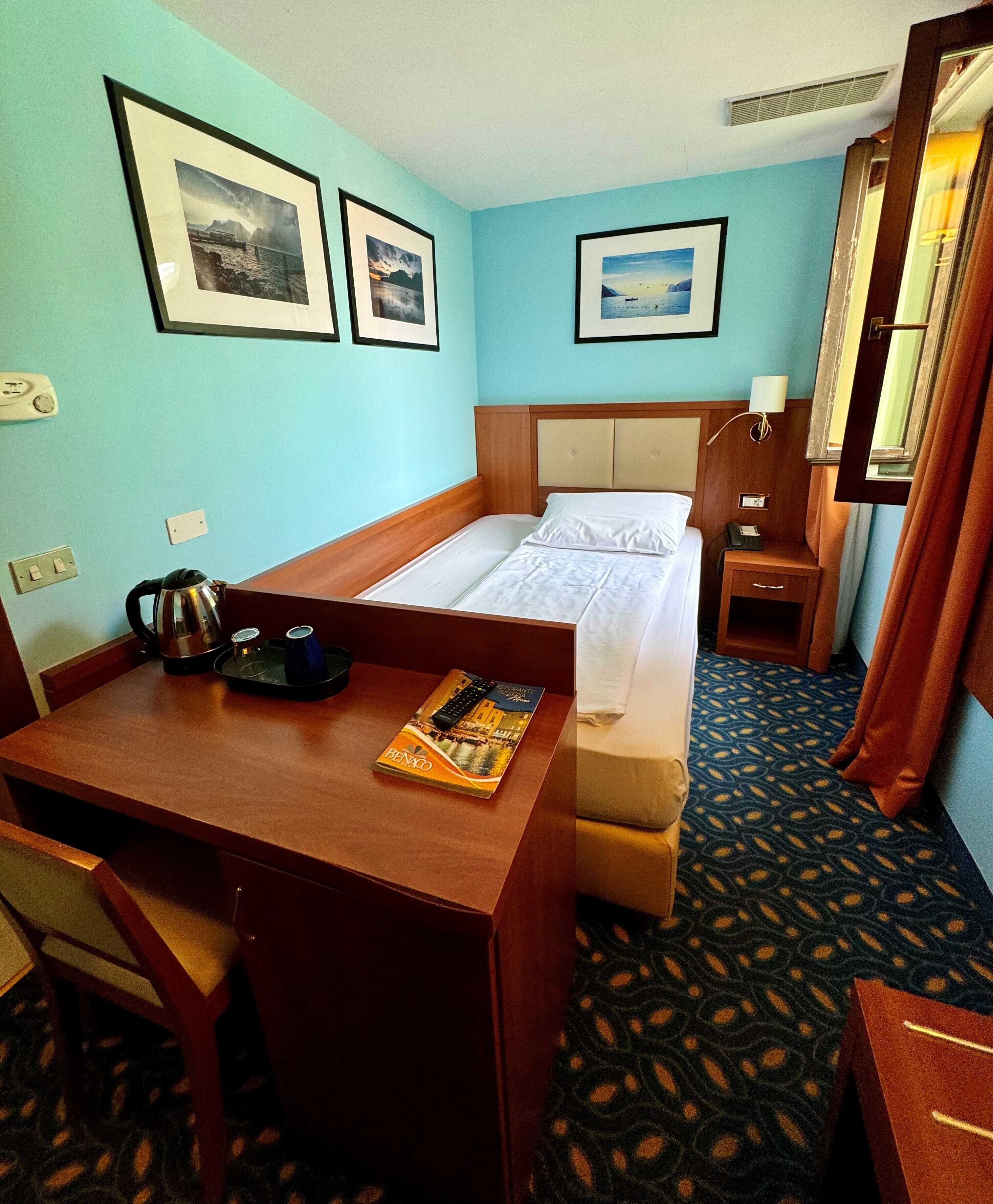 Una camera d'albergo con un letto, una scrivania, una sedia, un piccolo tavolo con un bollitore e un libro, un comodino con una lampada e quadri appesi alla parete.