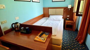 Una camera d'albergo con un letto, una scrivania, una sedia, un piccolo tavolo con un bollitore e un libro, un comodino con una lampada e quadri appesi alla parete.