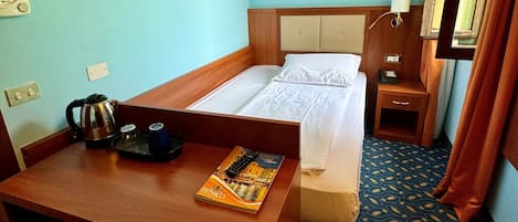 Hotelkamer met een bed, bureau, stoel, klein tafeltje met waterkoker en een boek, nachtkastje met lamp en ingelijste foto’s aan de muur.