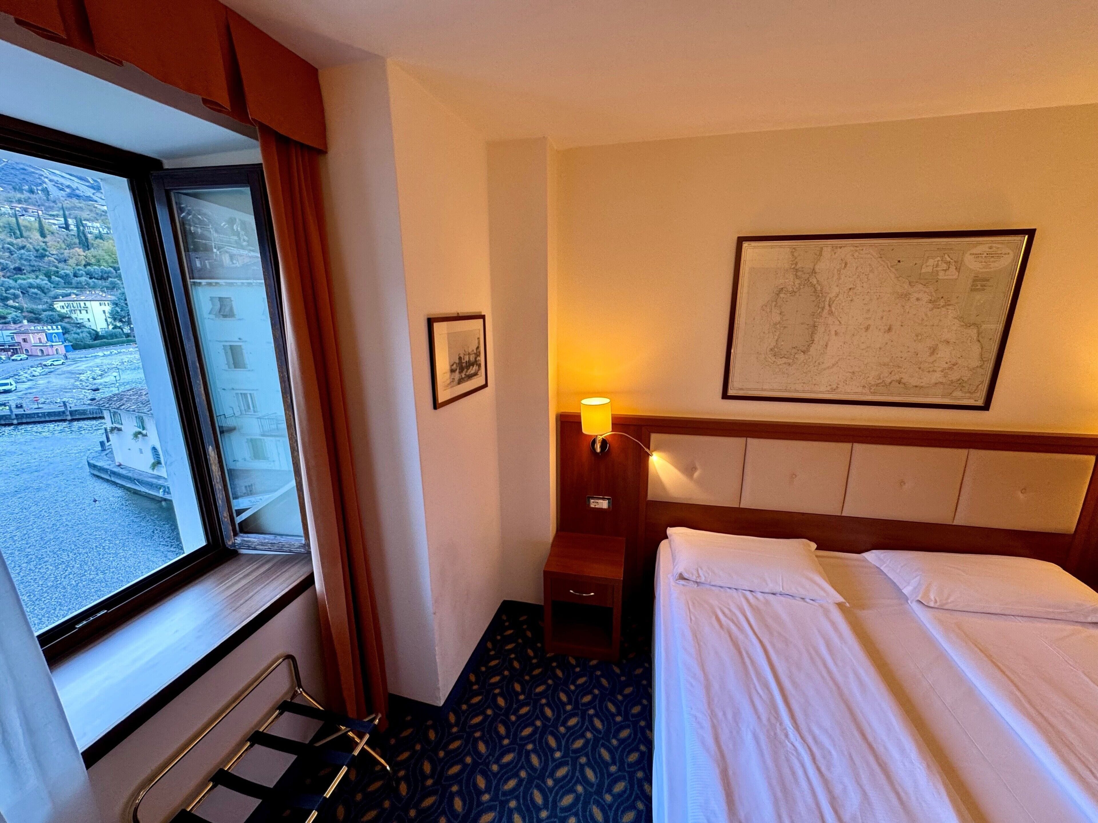 A hotel room with a bed, a nightstand, a lamp, a framed map on the wall, and a window with a view of a town.
