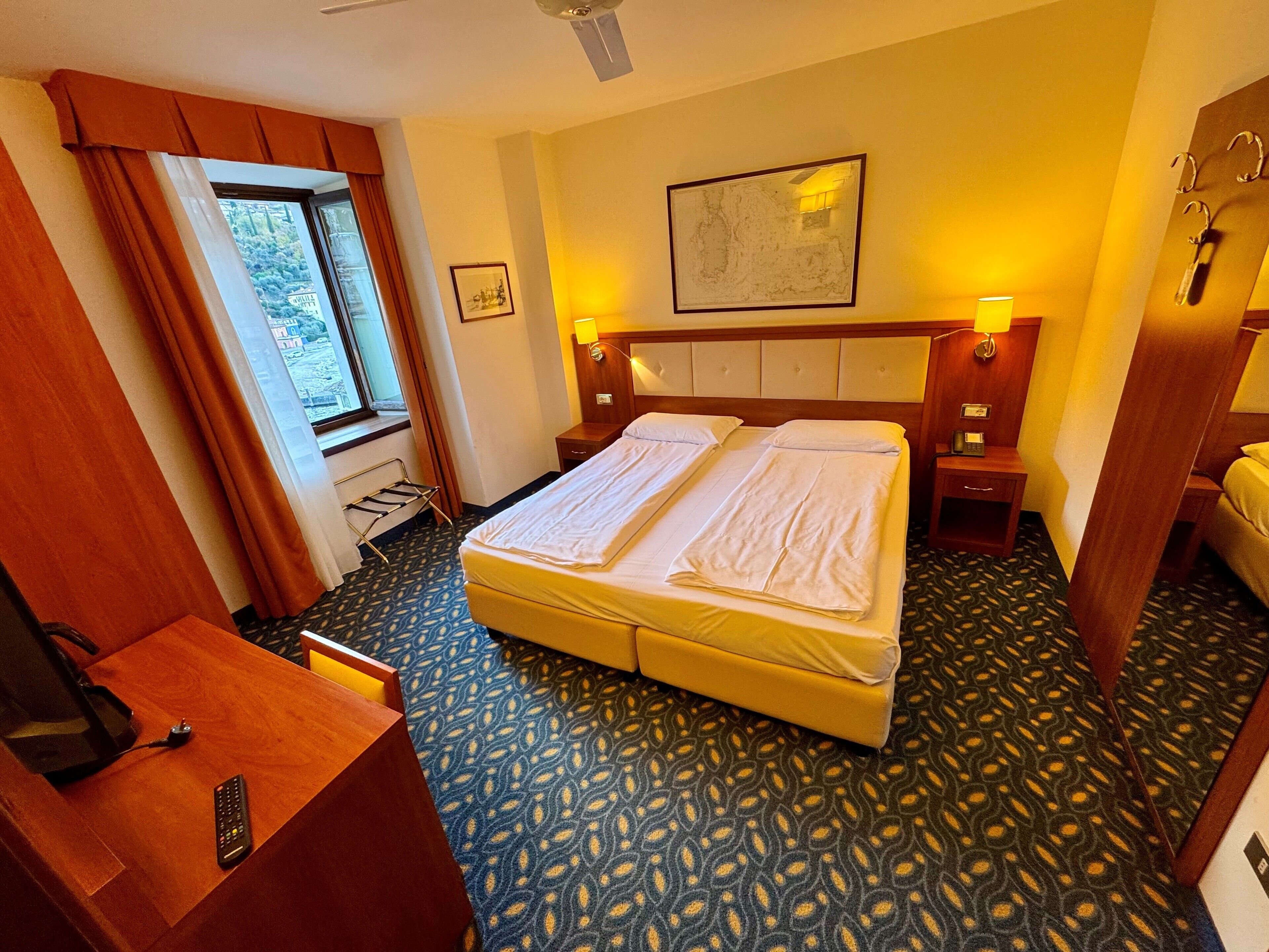 A hotel room with a double bed, two bedside tables with lamps, a desk with a telephone, and a window with curtains.