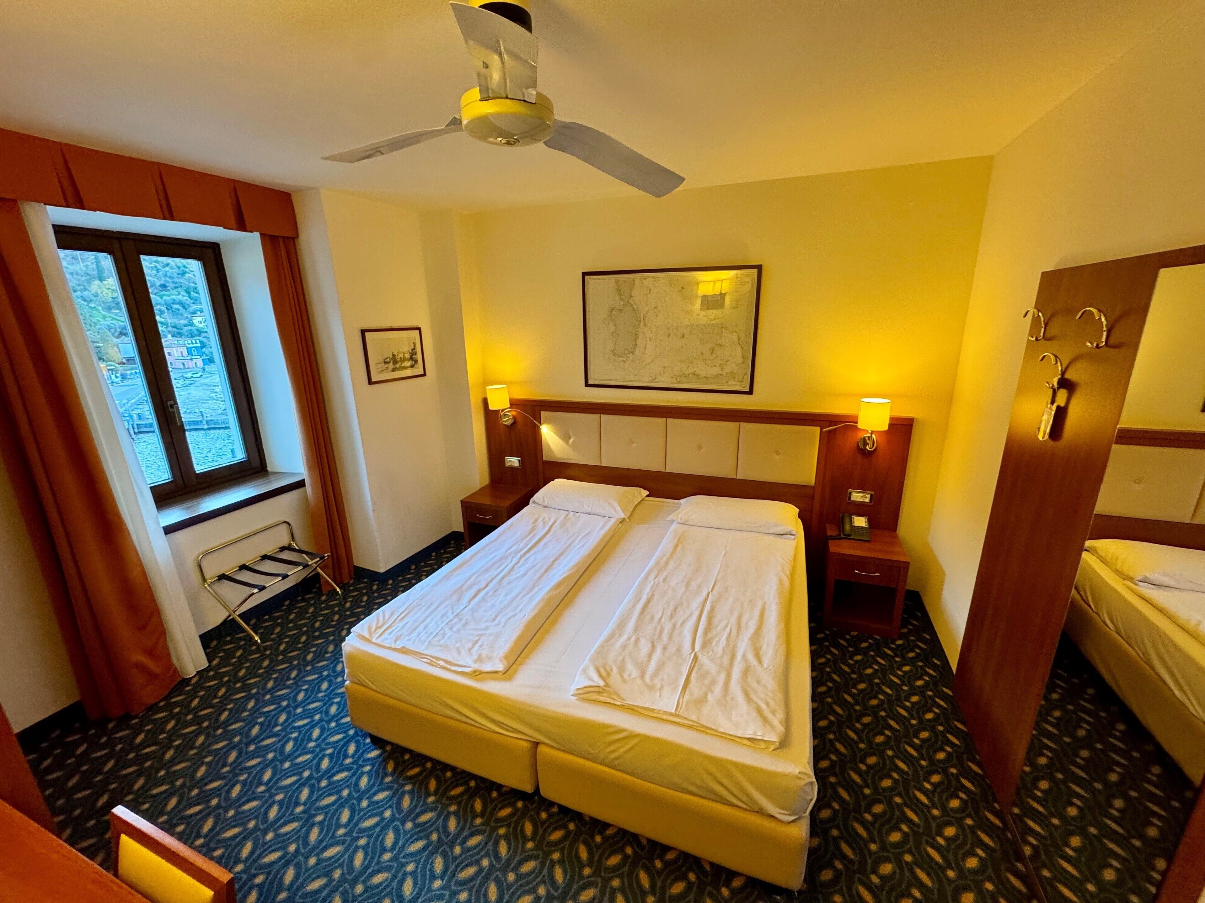A hotel room with a bed, bedside tables, a ceiling fan, a window with curtains, and a patterned carpet.