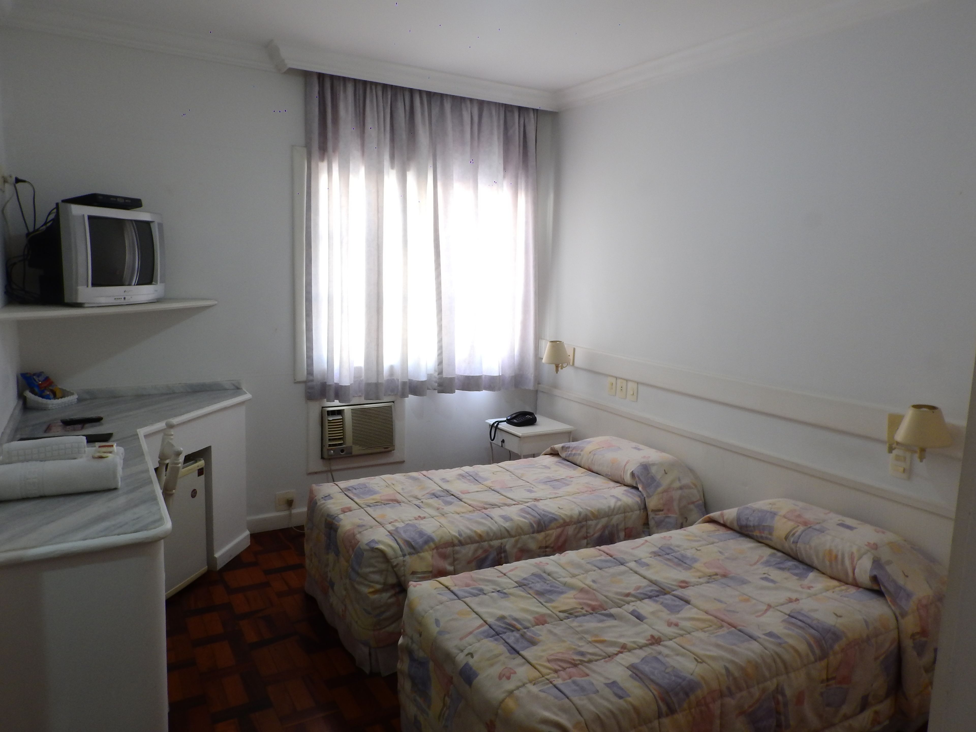 A hotel room with two beds, a TV on a shelf, a desk, and a window with curtains.