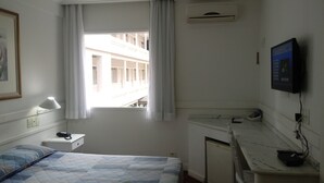 Habitación de hotel con cama, lámpara de noche, aire acondicionado, escritorio con computadora y ventana con cortinas.