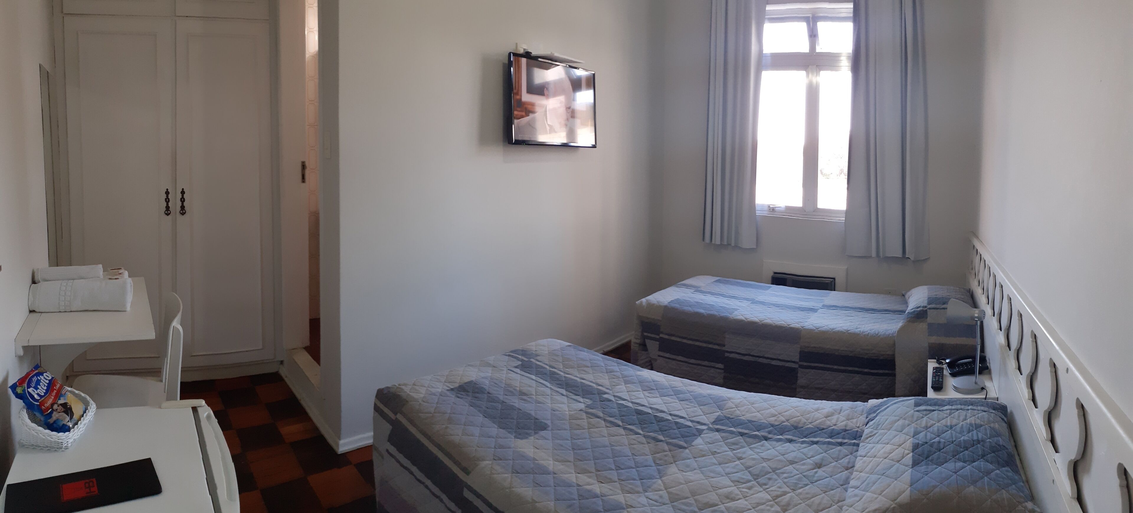 Un dormitori amb dos llits, una taula petita amb rellotge digital, un televisor penjat a la paret i una finestra amb cortines.