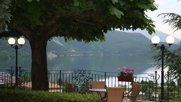 A terrace with wicker chairs and a table, overlooking a lake with mountains in the background. - Miralago (Molveno)