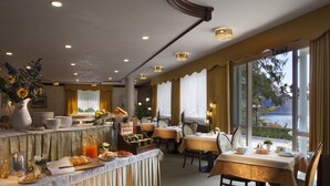 A hotel dining area with tables set for breakfast, a buffet with various food items, and large windows offering a view of the outdoors. - Miralago (Molveno)