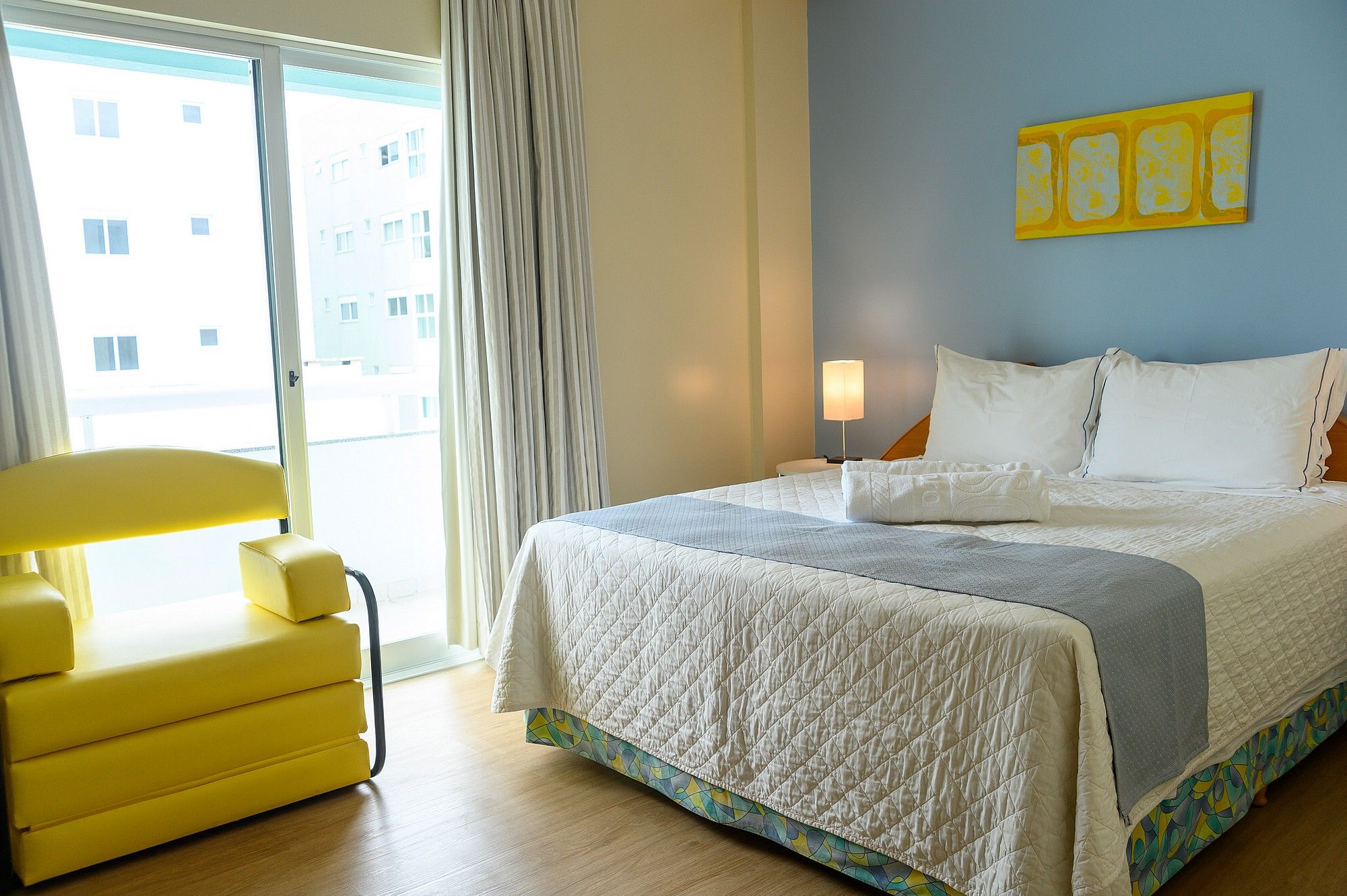 A hotel room with a bed, a chair, a lamp, and a view of buildings outside.