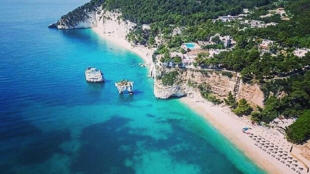 An aerial view of a coastal area with a beach, boats, and buildings on a hillside. - Hotel il Melograno (Mattinata)