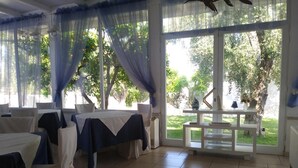 A room with tables set for dining, large windows with curtains, and a view of greenery outside. - Hotel il Melograno (Mattinata)