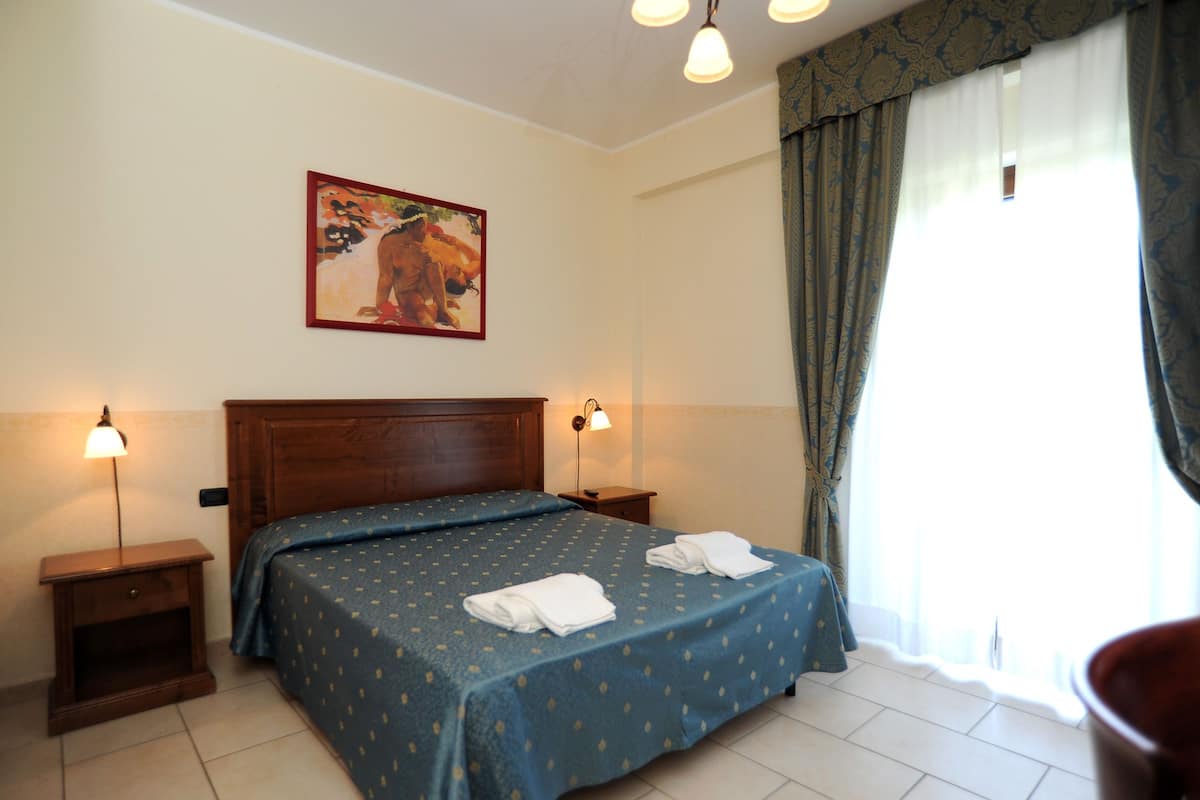 Una habitación de hotel con cama, dos lámparas de noche, un cuadro en la pared y una ventana con cortinas.