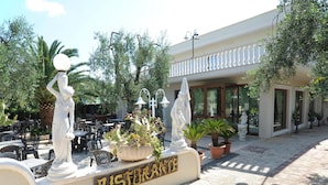An outdoor seating area with tables and chairs, a large planter with plants, and two white statues. - Hotel il Melograno (Mattinata)