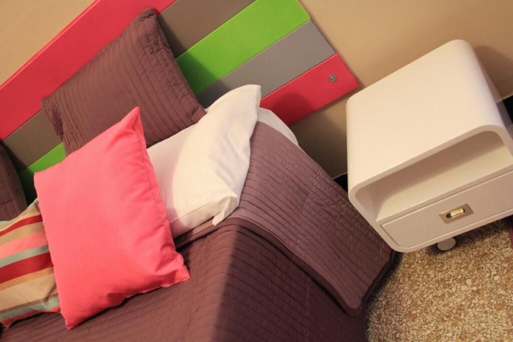Ein Bett mit braunem Kopfteil und Kissen in verschiedenen Farben, ein weißer Nachttisch und eine kleine, weiße Kommode.