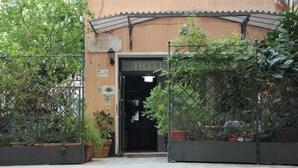 Entrada de hotel con un letrero que dice "HOTEL" sobre la puerta, rodeada de plantas en macetas y una valla metálica.