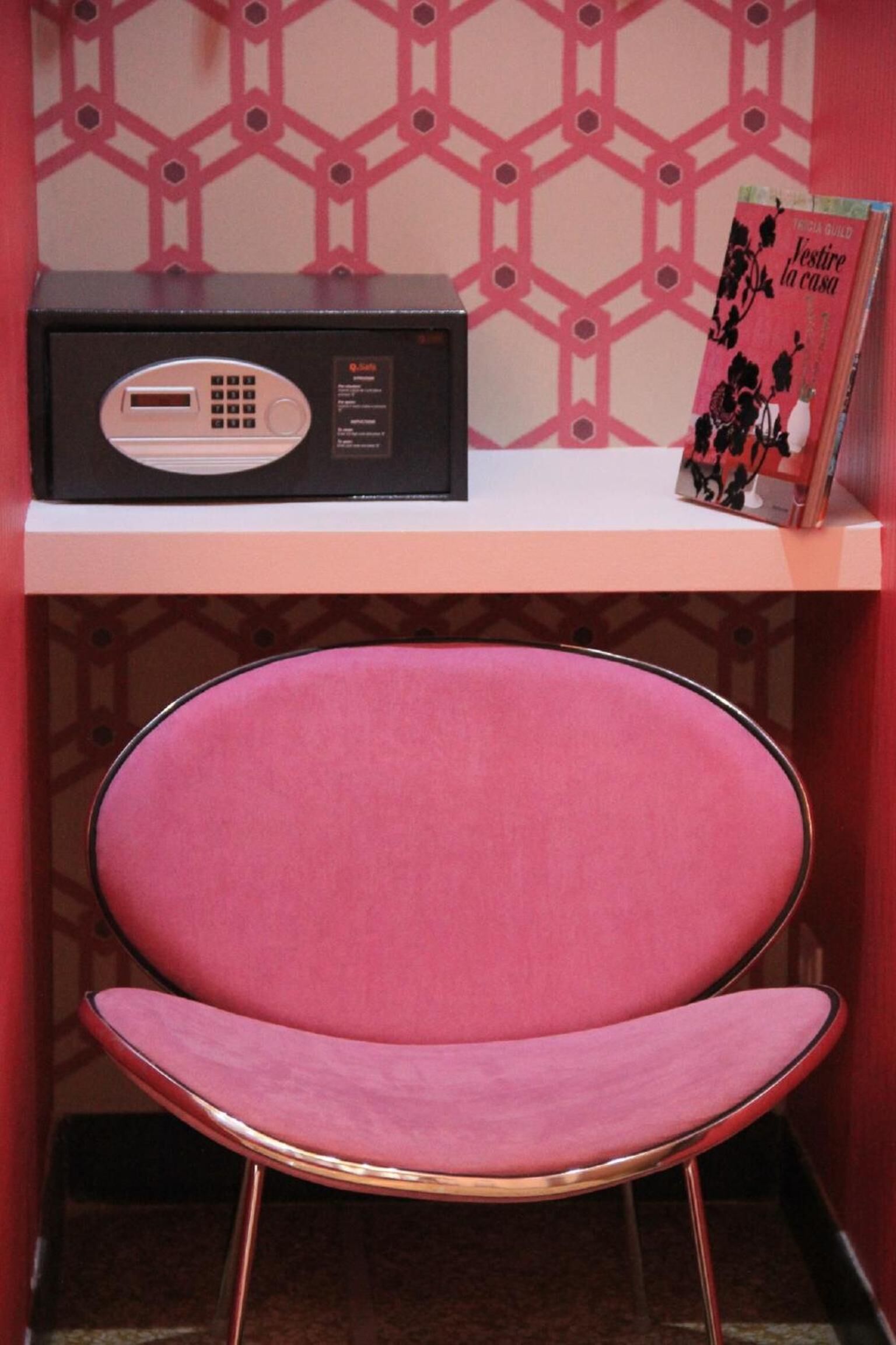 Una silla con estructura metálica y un cojín rosa, una radio negra, y un libro titulado 'Festine la casa' en un estante.
