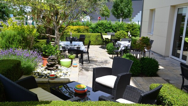 An outdoor patio with wicker furniture, stone tables, and a variety of plants. - Brit Hotel Saumur (Distre)