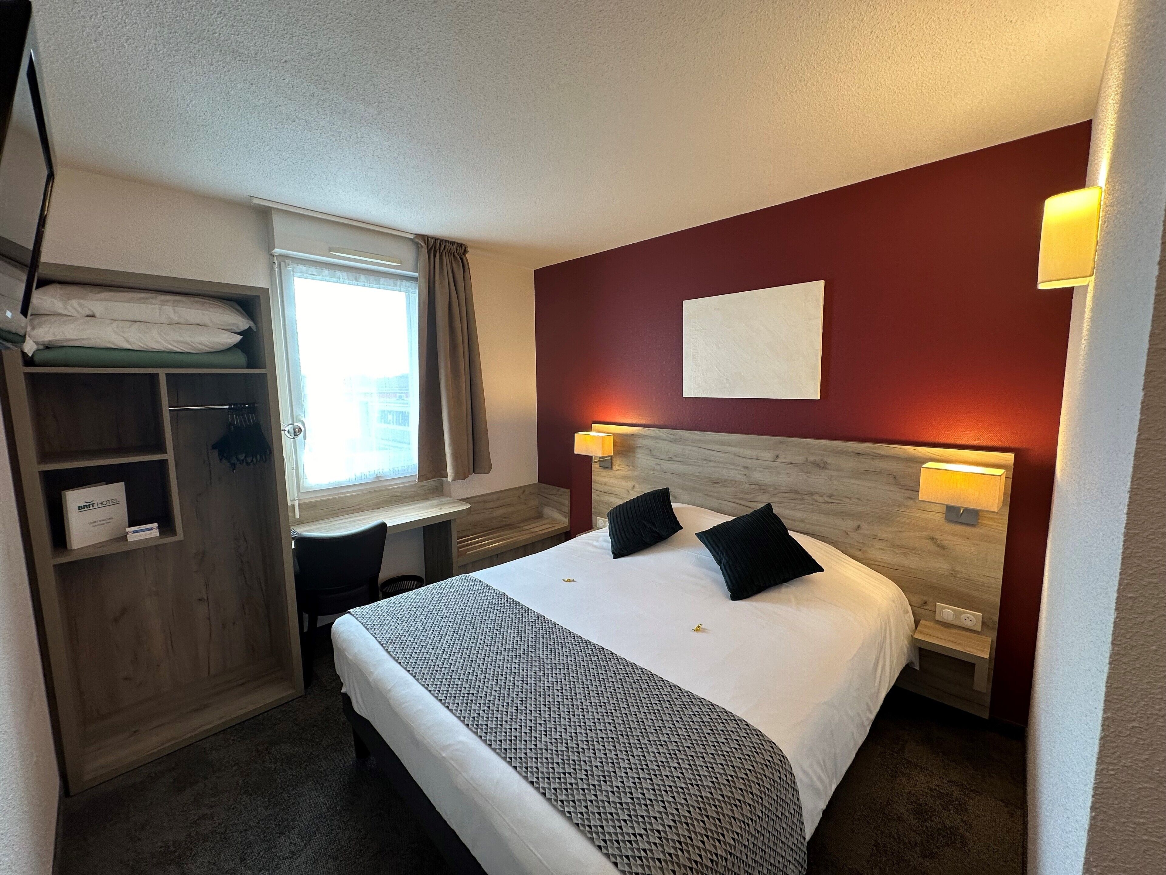 A hotel room with a bed, a desk, a wardrobe, and a window with curtains.