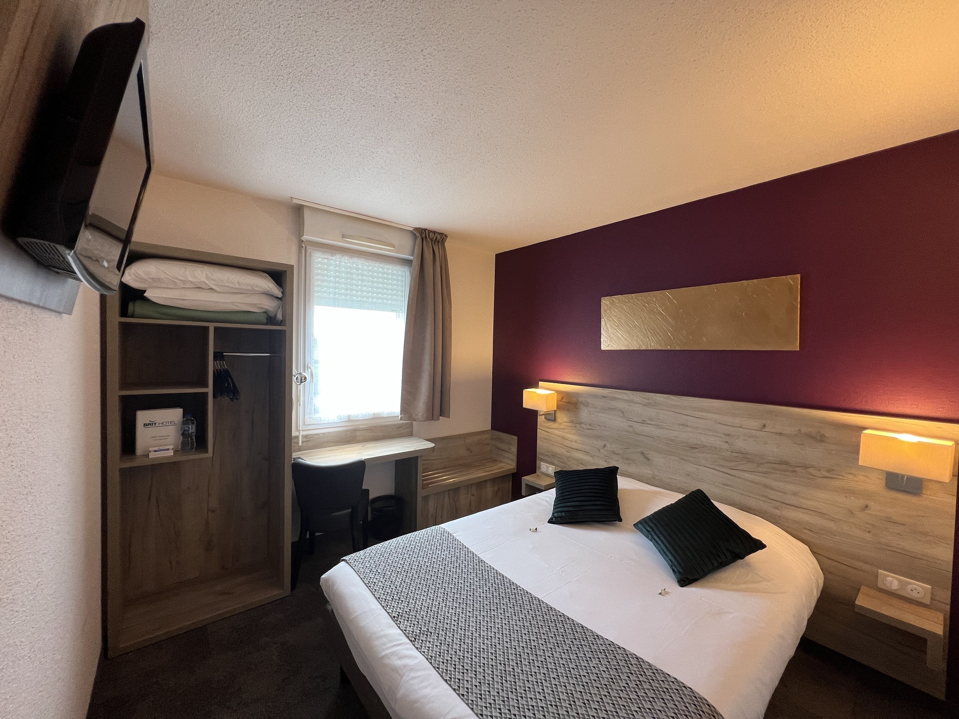 Habitación de hotel con cama, escritorio, armario, ventana con cortinas y televisor montado en la pared.