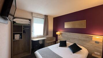 A hotel room with a bed, a desk, a wardrobe, a window with curtains, and a wall-mounted TV.