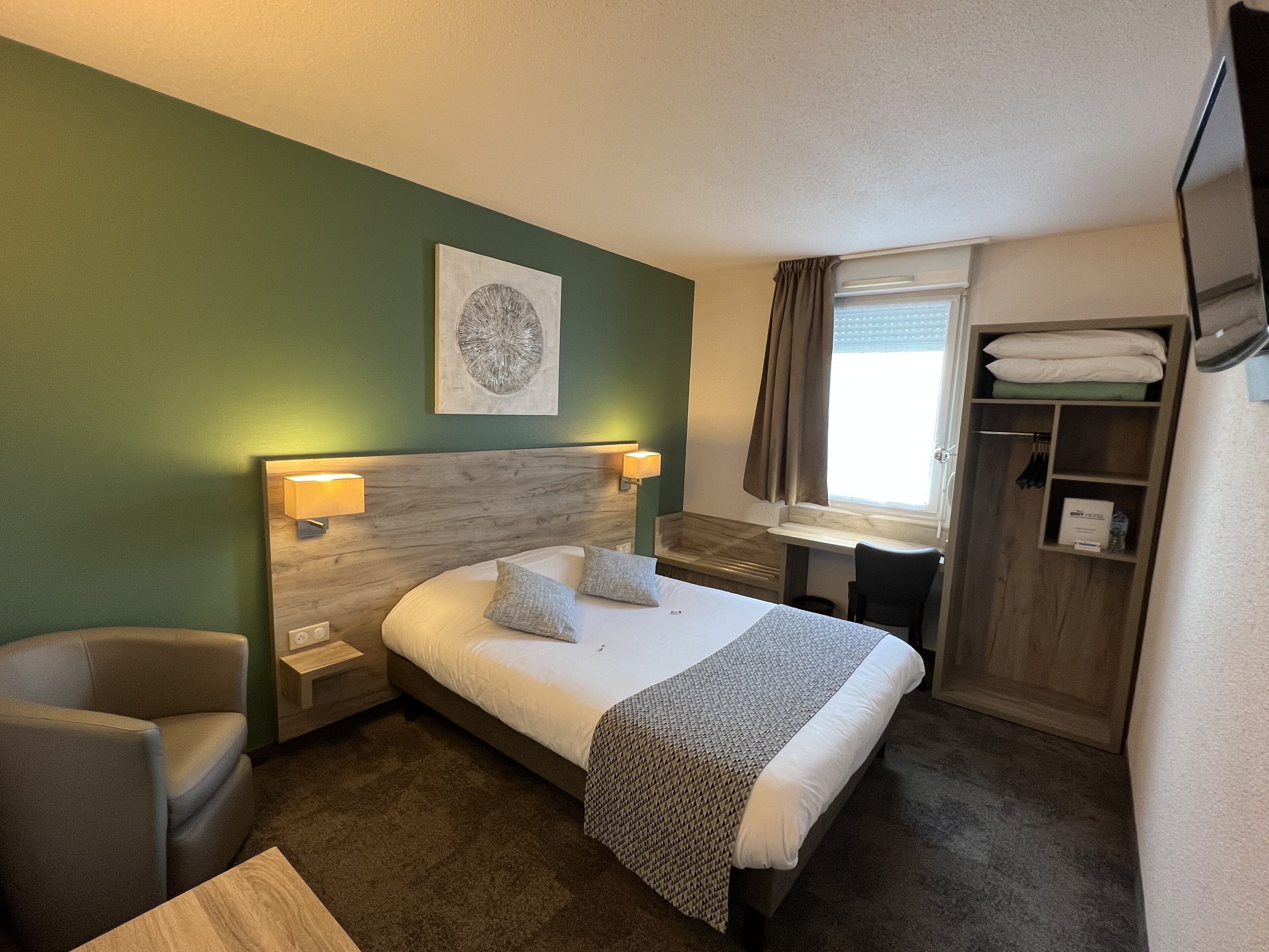 Una habitación de hotel con cama, dos lámparas de noche, cabecera, ventana con cortinas, armario y una pequeña mesa.