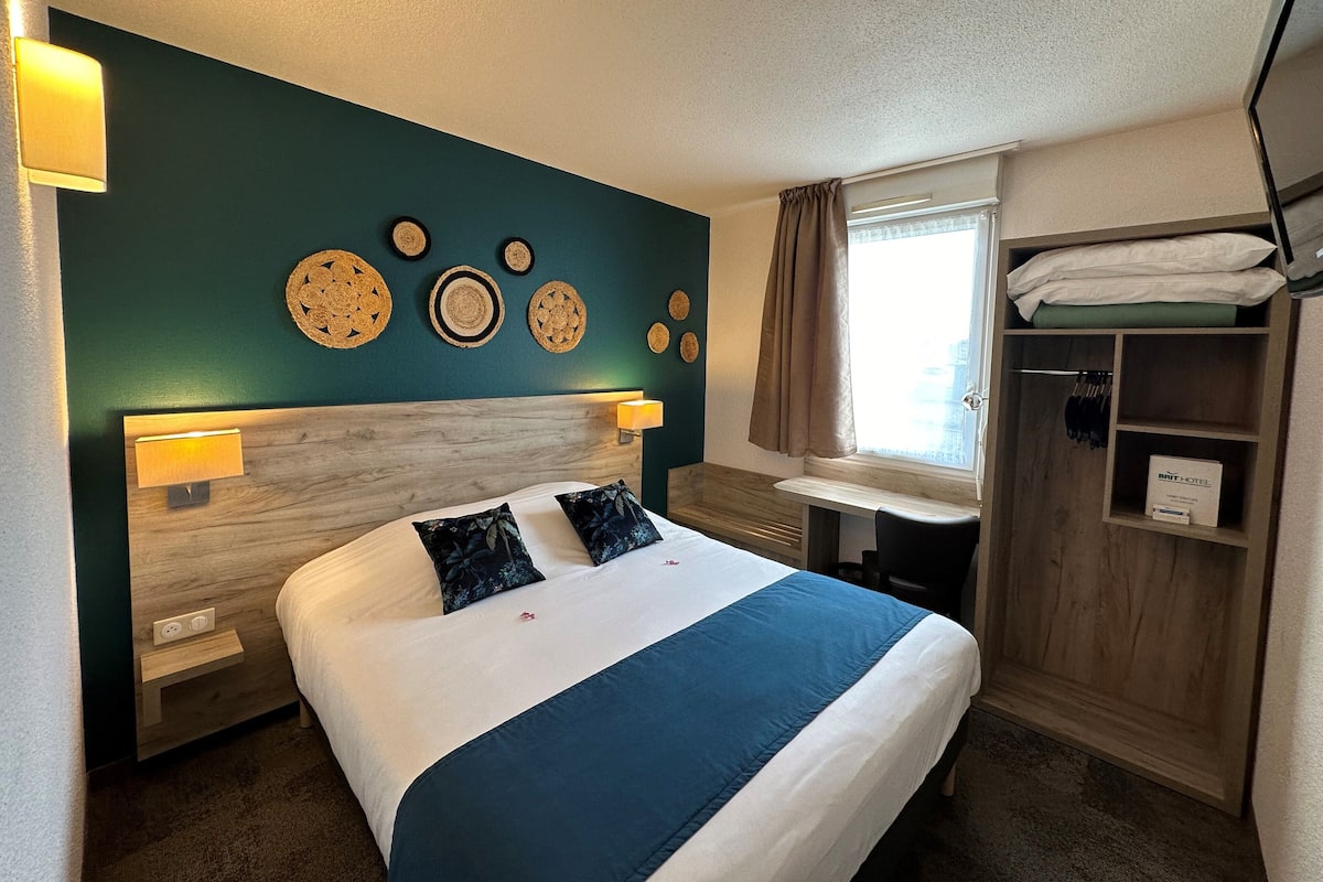 A hotel room with a bed, bedside lamps, a vanity with a mirror, a window with curtains, and a wardrobe.
