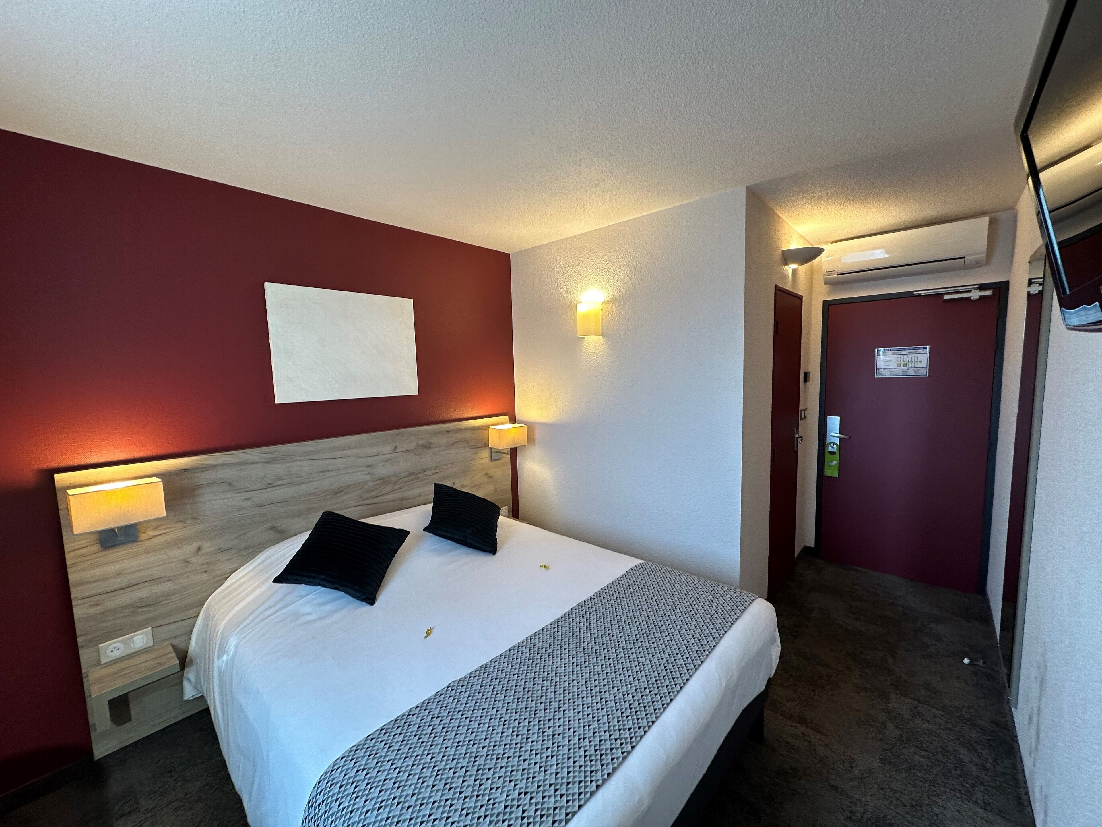 Habitación de hotel con cama, dos lámparas de noche, cabecero de madera, pared blanca y una puerta con un cartel.