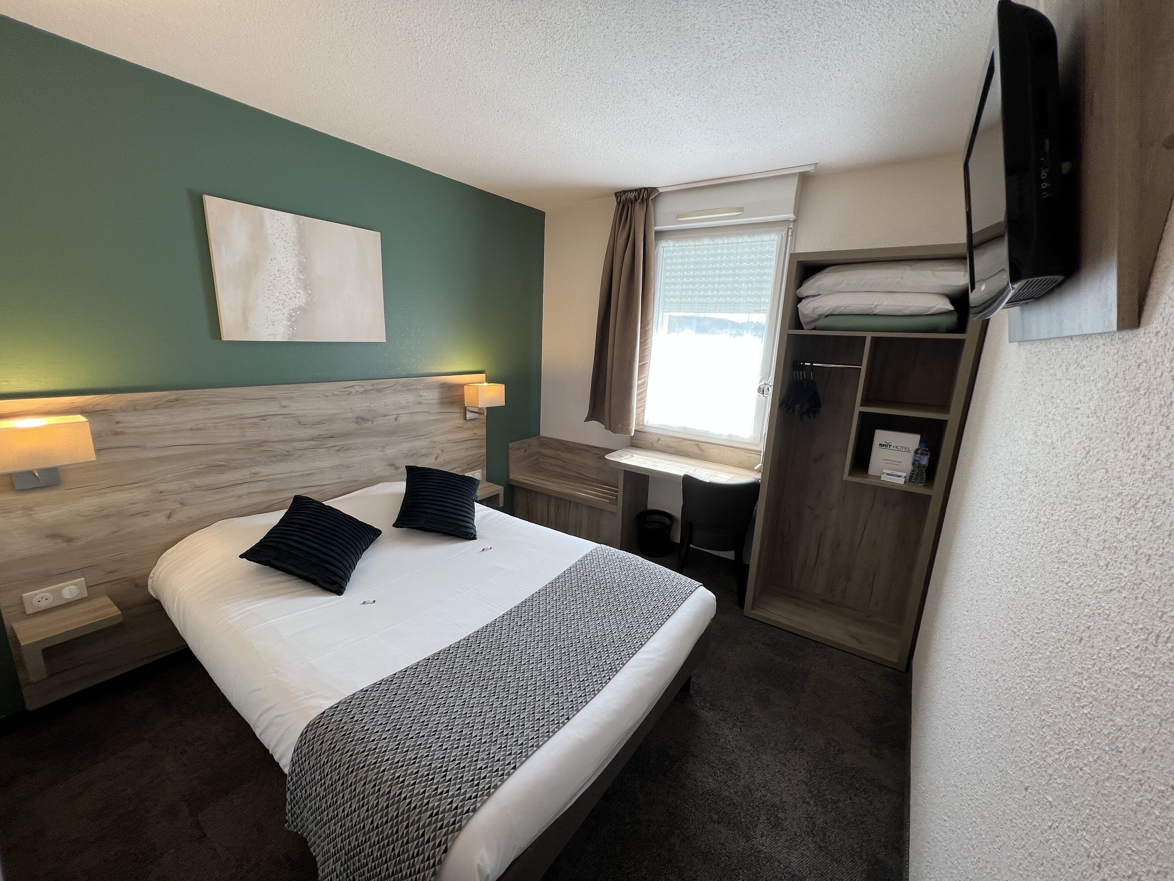 Habitación de hotel con cama, lámparas de noche, escritorio, silla, televisión y ventana con cortinas.