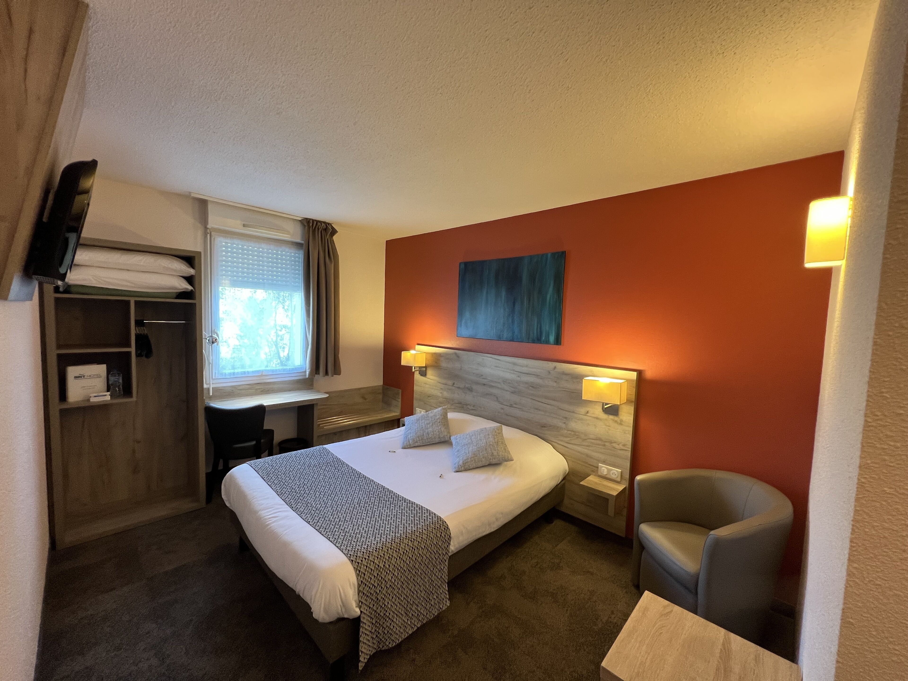 A hotel room with a bed, bedside tables, a chair, a desk, a wardrobe, and a window with curtains.