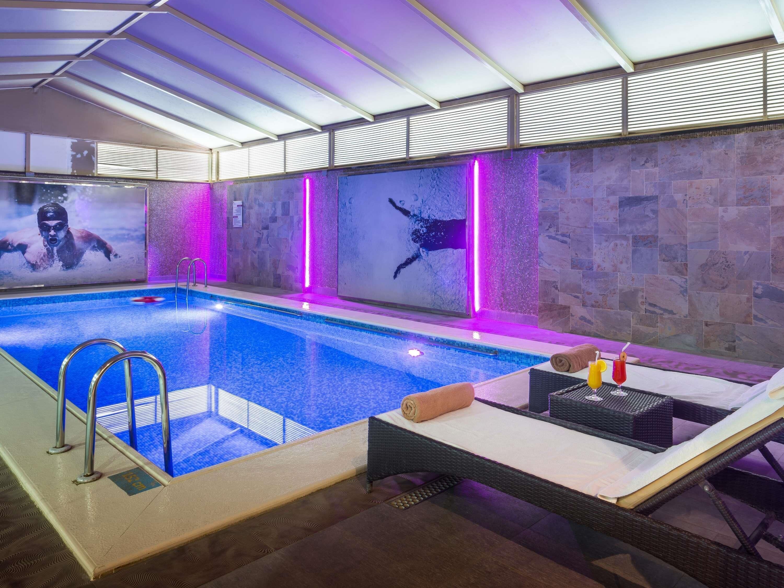 Une piscine intérieure séparée par une paroi en verre, un espace détente avec une table et des chaises, et un mur en pierre orné d’une fresque.