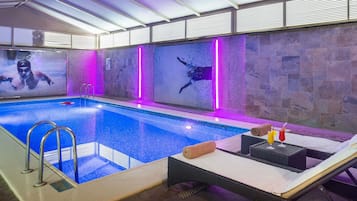 An indoor swimming pool with a glass barrier, a lounge area with a table and chairs, and a stone wall with a mural.