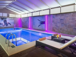An indoor swimming pool with a glass barrier, a lounge area with a table and chairs, and a stone wall with a mural. - Novotel Suites Riyadh Olaya (Riyadh)