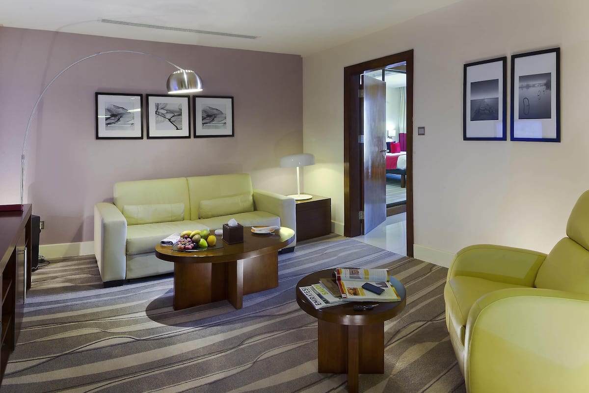 Quarto de hotel moderno com sofá, mesa de centro e uma poltrona amarela. Há uma luminária, uma pequena mesa com revistas e uma porta que dá para outro cômodo.