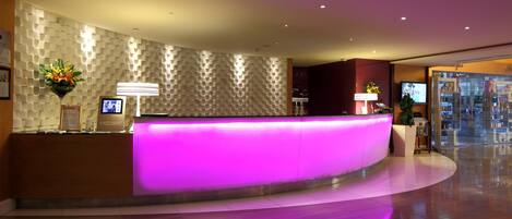 Un'accogliente area reception di un hotel moderno, con una scrivania a forma di arco, uno schermo digitale e un muro con motivo decorativo.