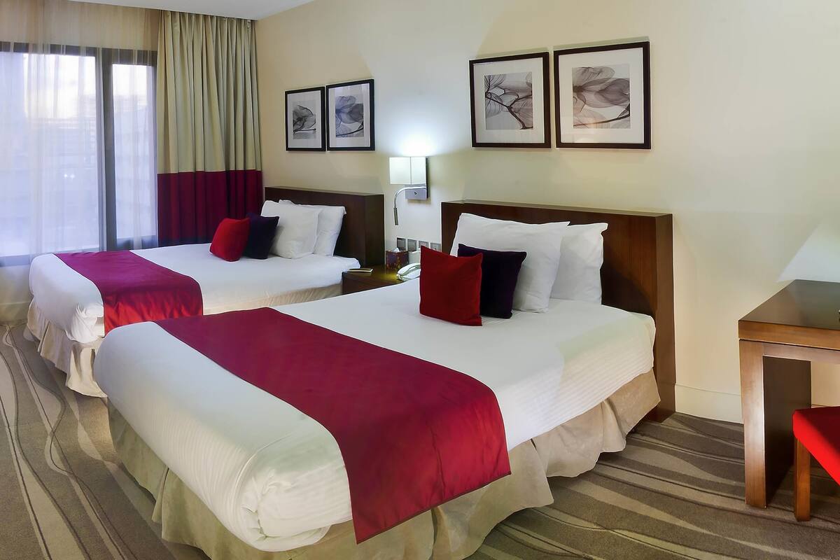 Quarto de hotel com duas camas, uma mesa de cabeceira, abajur e obras de arte na parede.