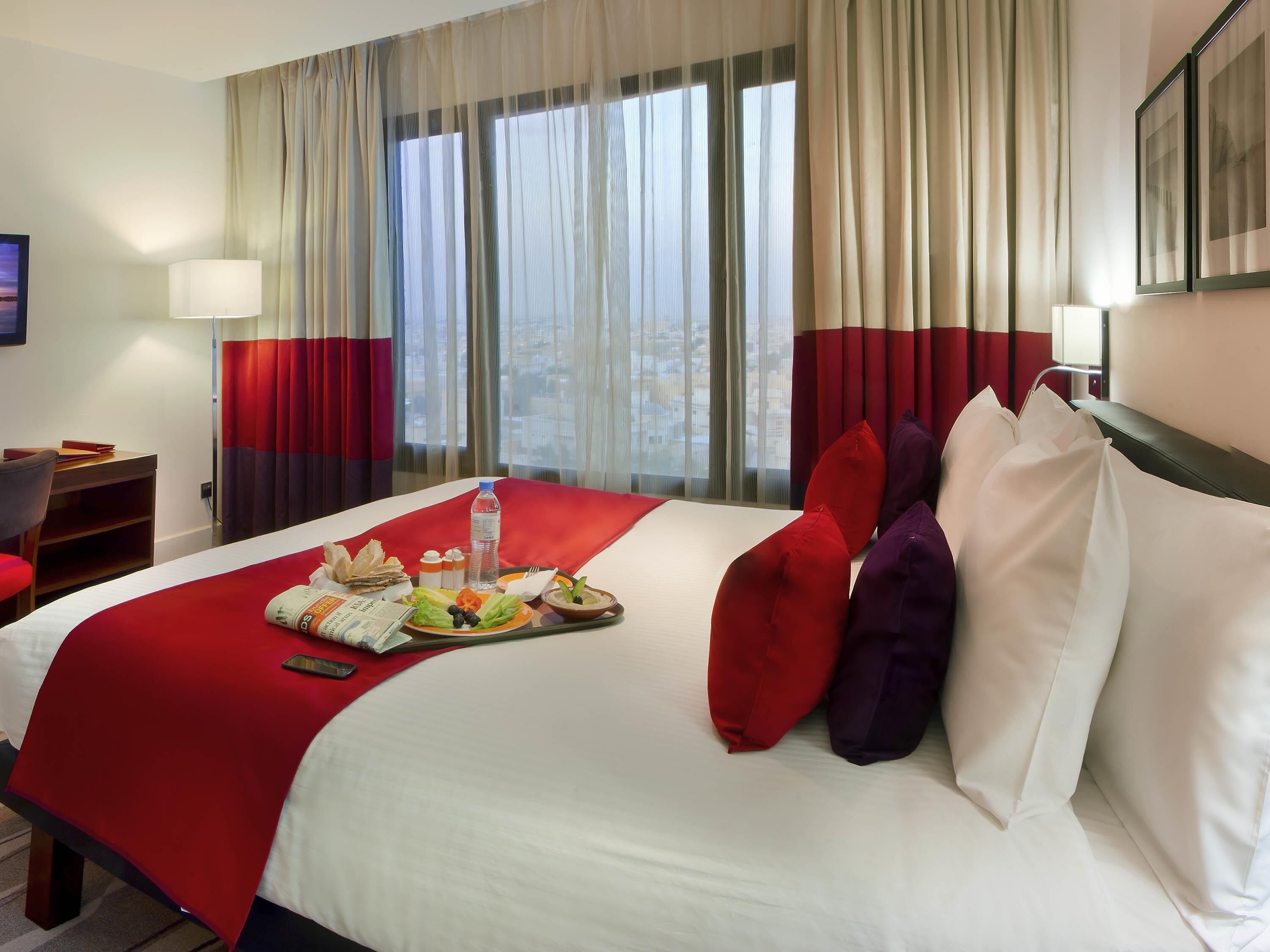 A hotel room with a bed, a desk, a chair, a window with curtains, and a tray with breakfast items.