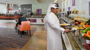 Uma área de buffet com diversas opções de comida, um balcão com alguém servindo e uma área de estar com carpete estampado.