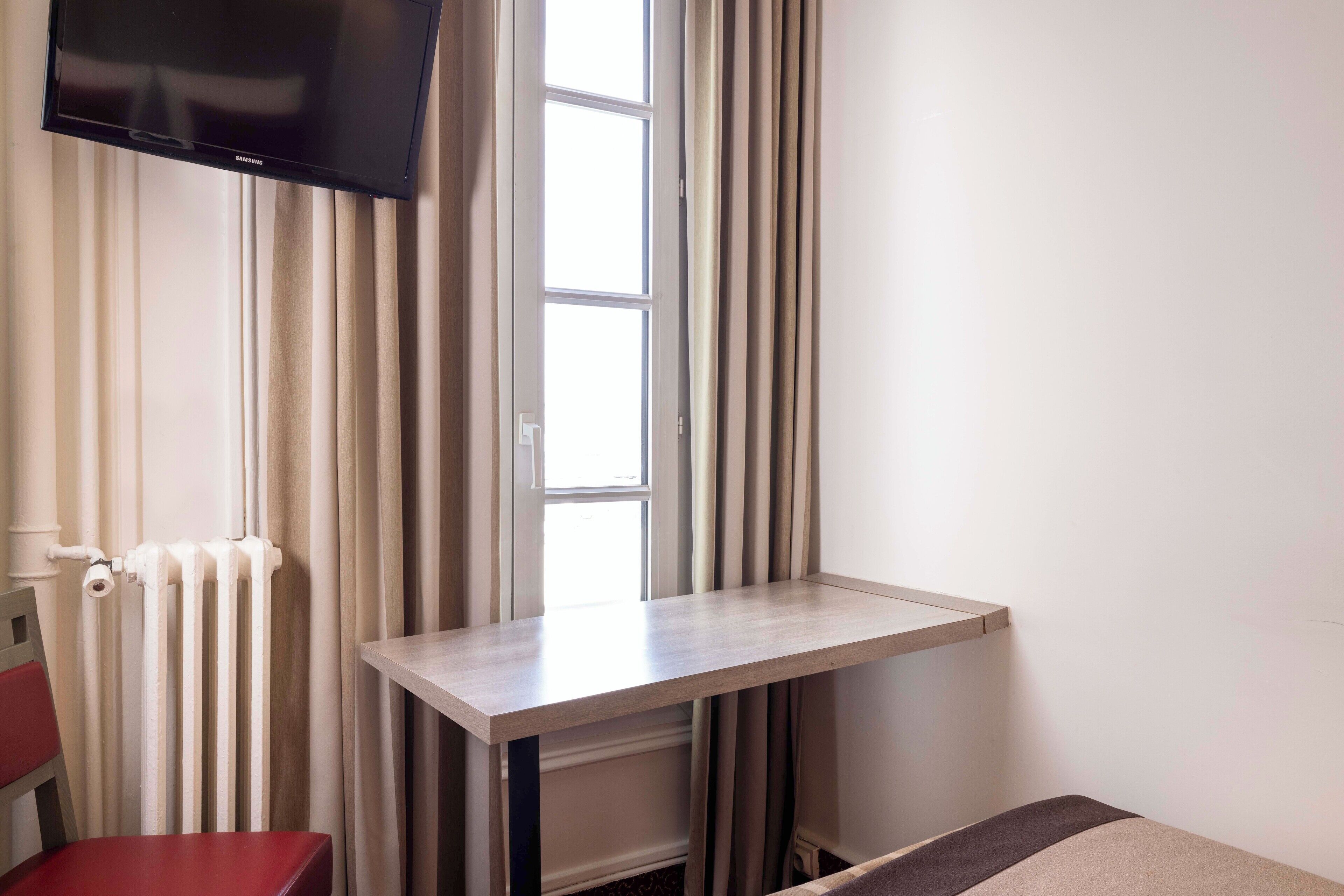 A room with a bed, a small table, a TV mounted on the wall, a radiator, and a window with curtains.