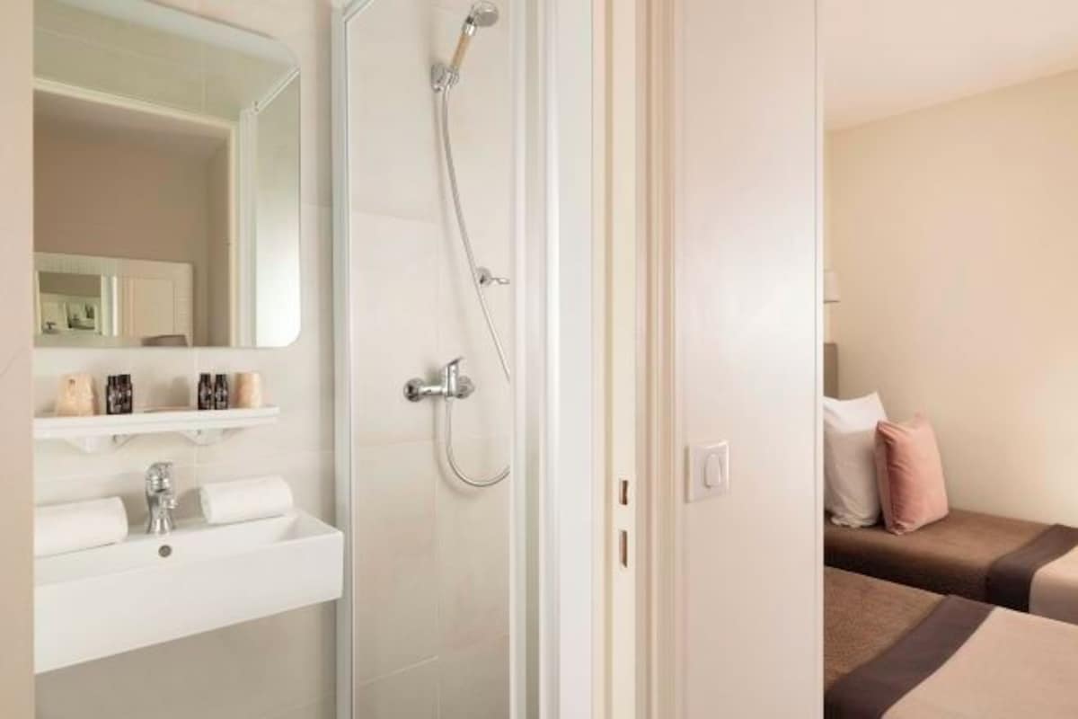 A hotel room with a bathroom featuring a sink, mirror, and shower. There is a bed with pillows and a bedside table.