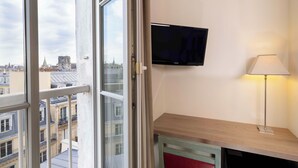 A room with a TV mounted on the wall, a lamp, and a view of the cityscape through a window. - Hotel Saint Pierre (Paris)