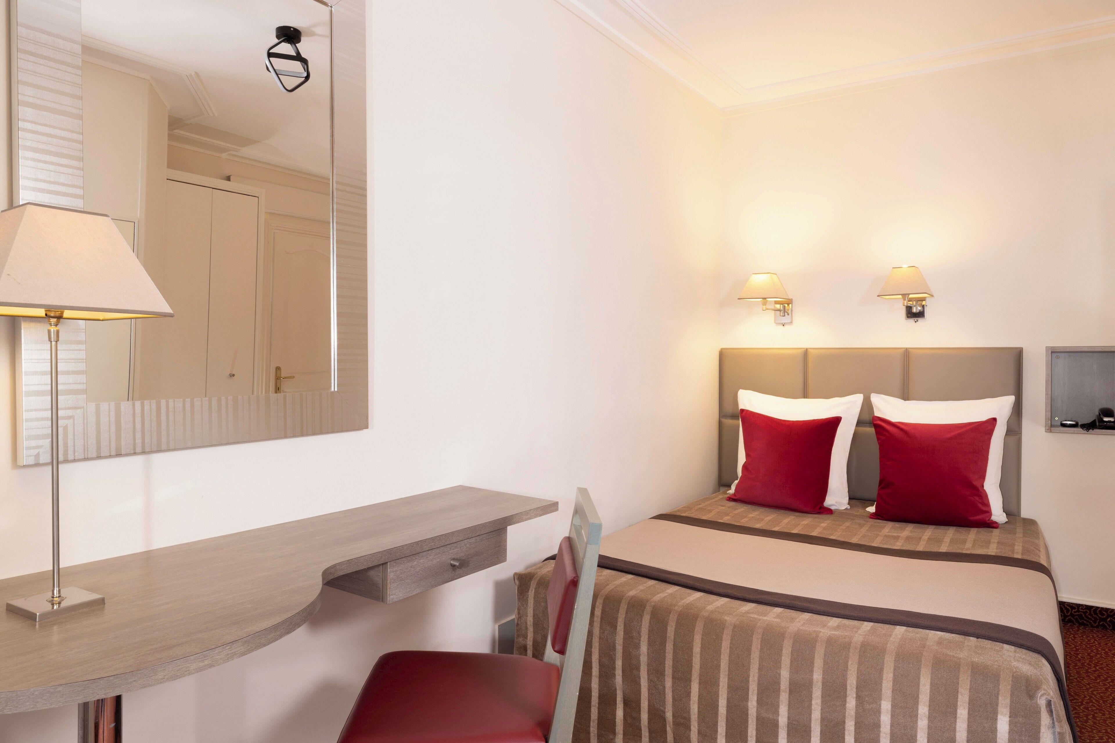 A hotel room with a bed, a desk, a chair, a mirror, and two wall-mounted lamps.