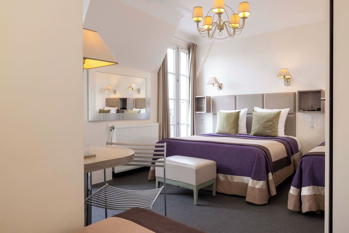 A hotel room with a bed, bedside tables, a chair, a small table, and a chandelier.