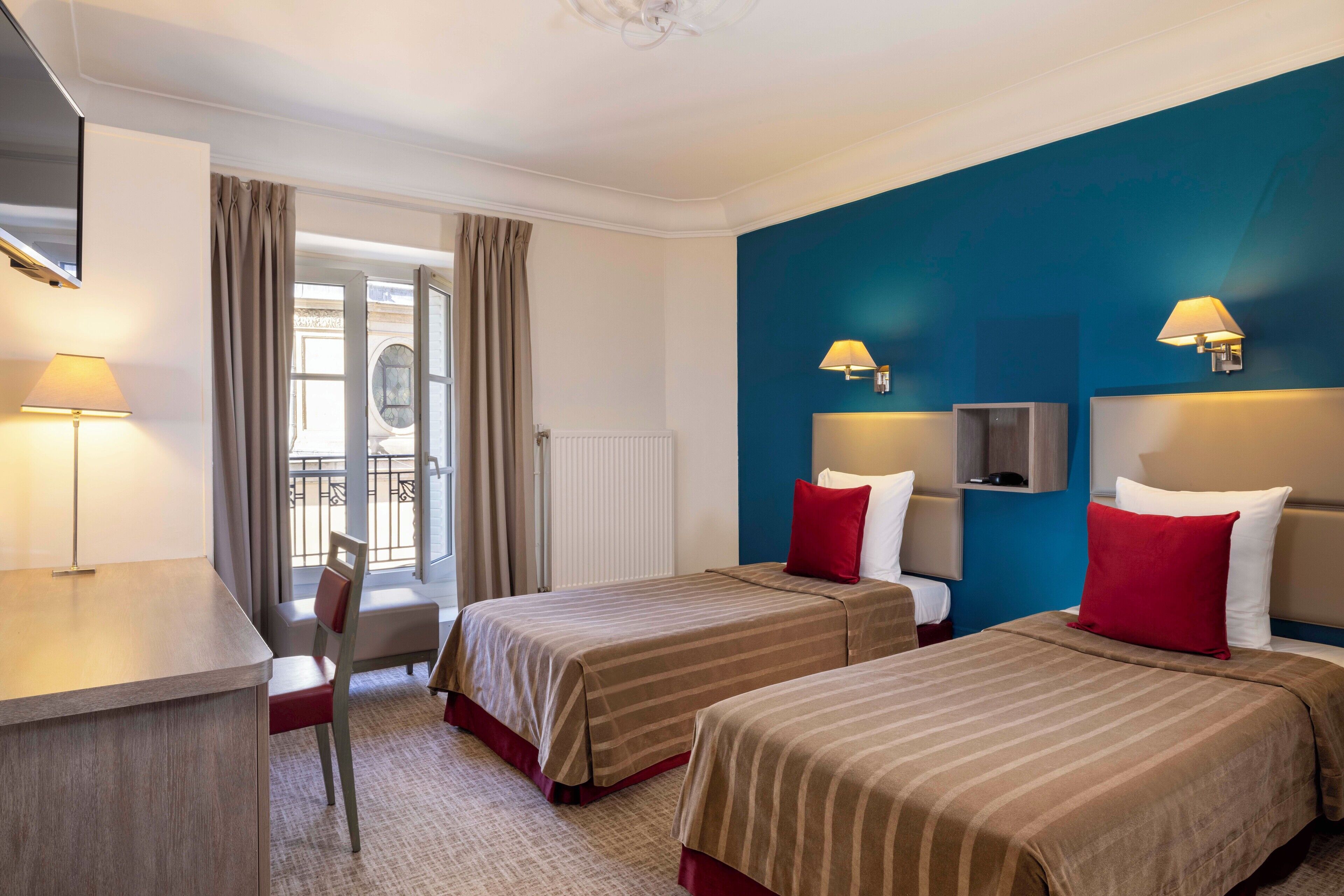 Habitación de hotel con dos camas, un escritorio y una silla. Cuenta con dos lámparas de noche y vistas al exterior a través de una ventana con cortinas.