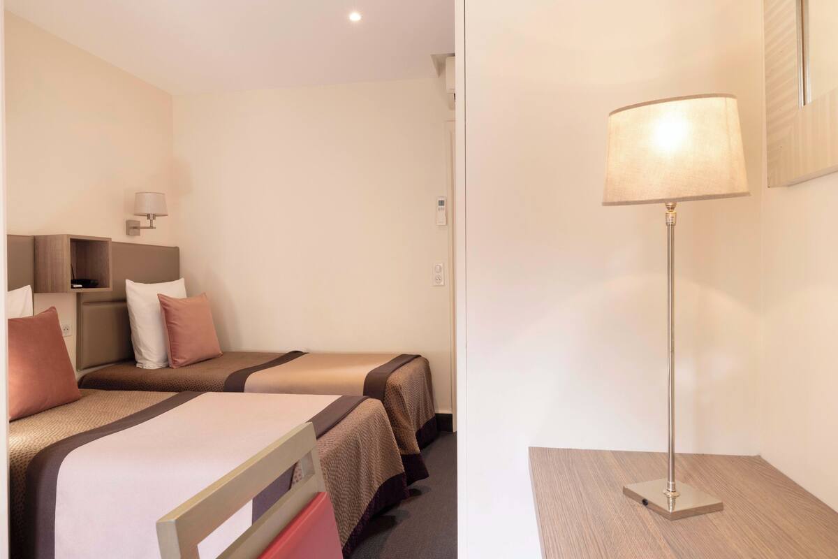 A hotel room with two beds, a bedside table, a lamp, and a wall-mounted light fixture.