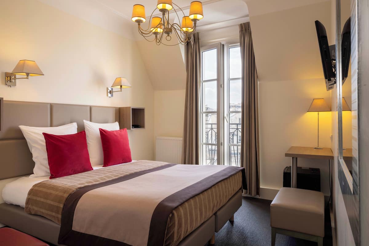 A hotel room with a large bed, two red pillows, a chandelier, a desk with a lamp, and a view of the city through the window.