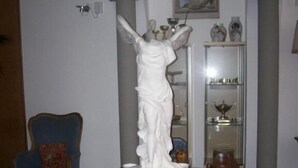 Une statue représentant une femme tenant une torche, placée sur une table en bois dans une pièce où se trouvent un fauteuil bleu et une vitrine contenant des objets décoratifs.