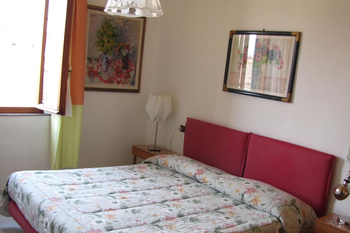 A bedroom with a floral bedspread, a red headboard, a hanging light fixture, a framed picture on the wall, and a window with curtains.