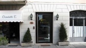 Une façade d’immeuble avec une enseigne indiquant « Ciadelli E. » et une porte au-dessus de laquelle est fixée une autre enseigne. Deux plantes en pot sont placées sur le trottoir, devant la porte.