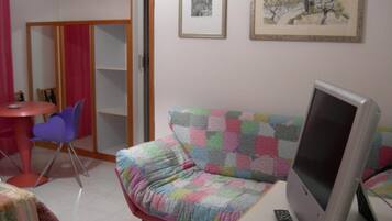 Una habitación pequeña con un sofá acolchado de colores, un televisor, un control remoto, una mesita con una silla y una puerta que da a otra habitación.