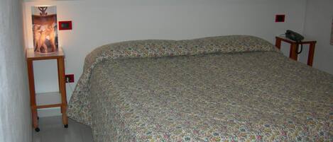 Un dormitorio con colcha de estampado floral, una lámpara de noche, una mesita con teléfono y un espejo en la pared.
